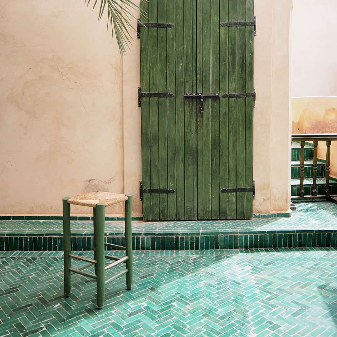 Rustic courtyard with green herringbone tile floor, simple wooden stool, tall green door set in weathered beige plaster wall, tiled steps, and palm frond adding Mediterranean-inspired natural charm.