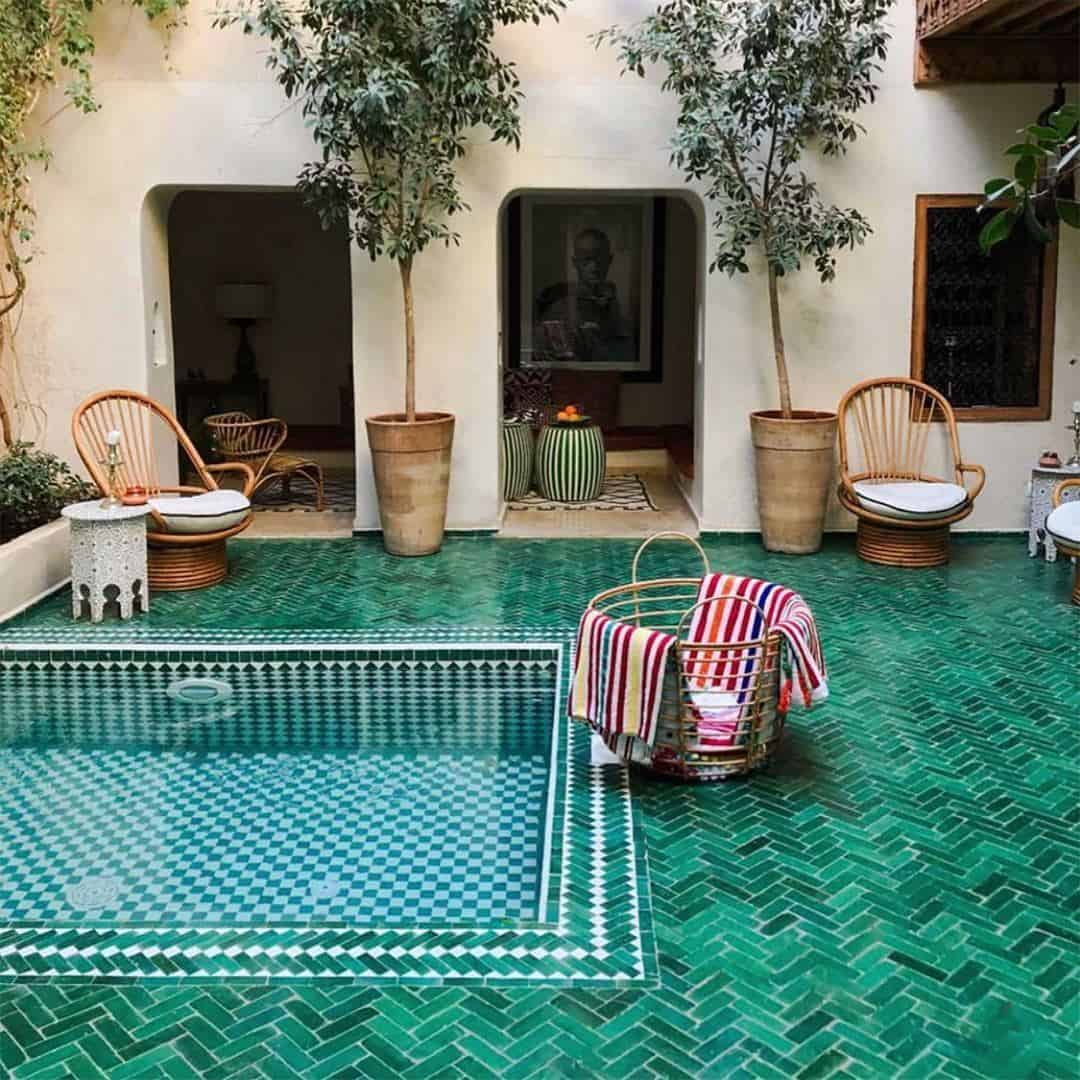 Courtyard with small rectangular pool tiled in green and white geometric pattern, surrounded by green herringbone floor, wicker chairs, potted trees, striped towels, and arched doorways creating serene Mediterranean-inspired atmosphere.