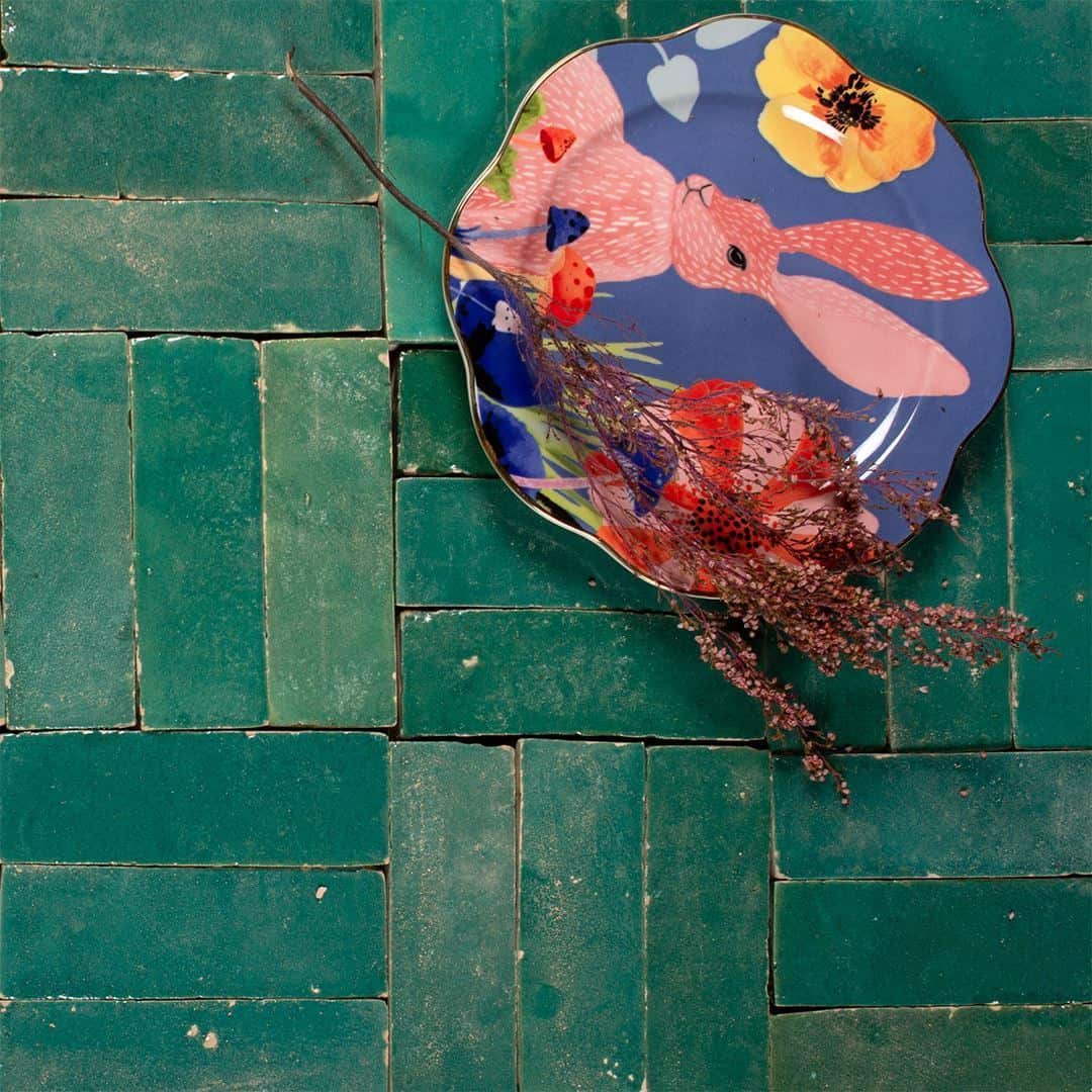 Decorative scalloped plate with colorful design featuring pink rabbit, flowers, and blue accents, resting on green herringbone tile surface with dried foliage sprig adding texture and contrast.