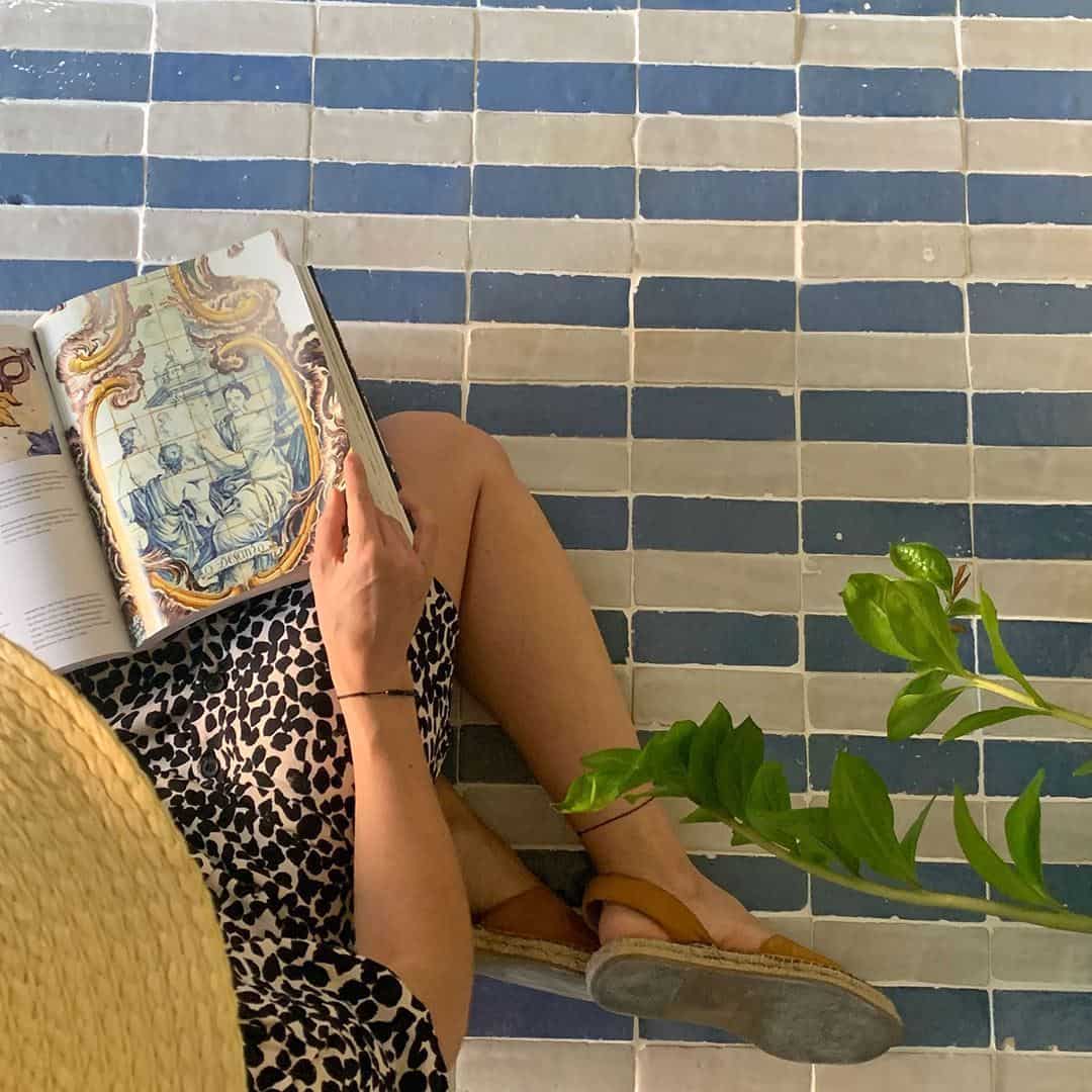 Person in patterned dress, sandals, and straw hat sitting on blue and gray tiled floor, holding open book with ornate illustration, next to leafy plant