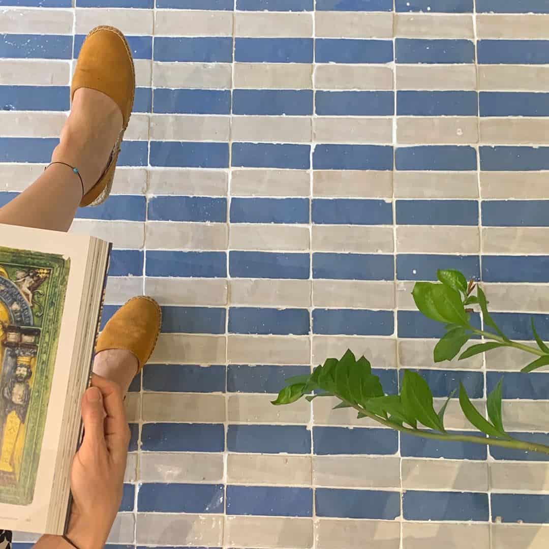 Person in sandals sitting on blue and gray tiled floor, holding open book with ornate illustration, next to leafy plant