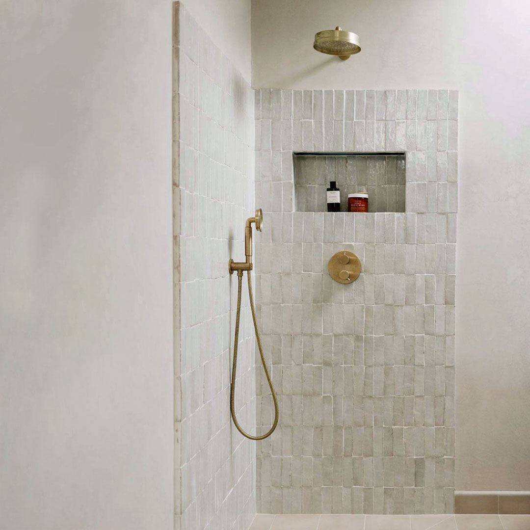 Minimalist shower area with light beige square tiles, a brass showerhead and handle, and a built-in recessed shelf holding two bottles. 