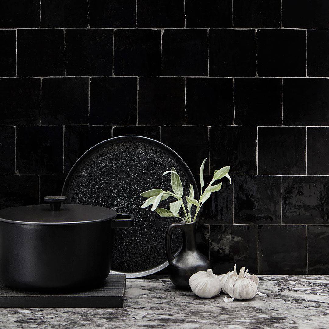 Modern kitchen scene with matte black cooking pot on trivet, speckled black plate against glossy tiled wall, ceramic pitcher with herbs, and garlic bulbs on marble countertop, creating minimalist elegance.