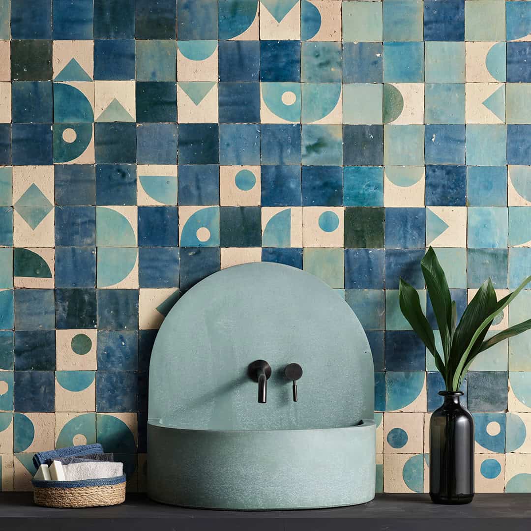 Wall tiles in blue and beige geometric patterned tiles, and a black vase with green leaves sits beside the sink.