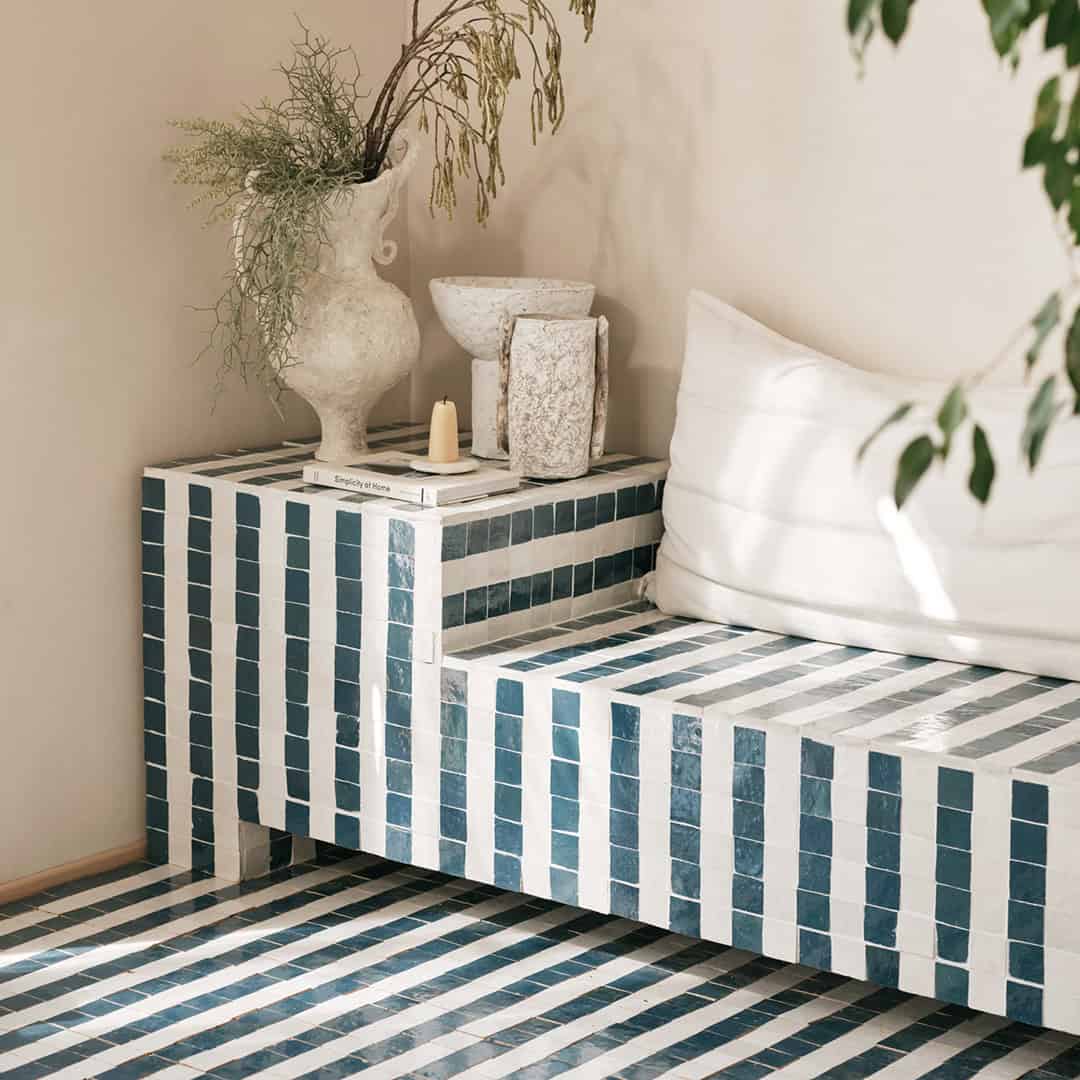 Modern corner bench covered in teal and white striped tiles, decorated with white cushion, textured vase with branches, stone vessels, candle, and book titled “Simplicity of Home.
