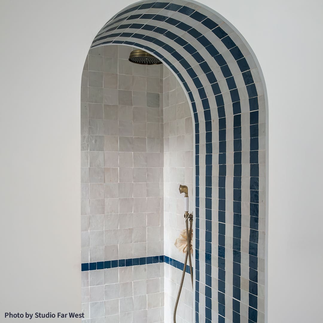 Arched shower alcove with light gray tiled walls and alternating dark blue and gray striped arch, featuring brass ceiling showerhead, handheld fixture, and sponge, creating modern geometric bathroom design.