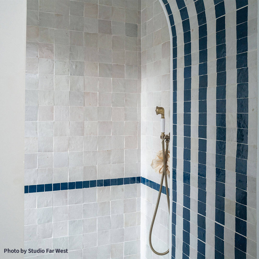 Arched shower alcove with light gray tiled walls and alternating dark blue and gray striped arch, featuring brass ceiling showerhead, handheld fixture, and sponge, creating modern geometric bathroom design.