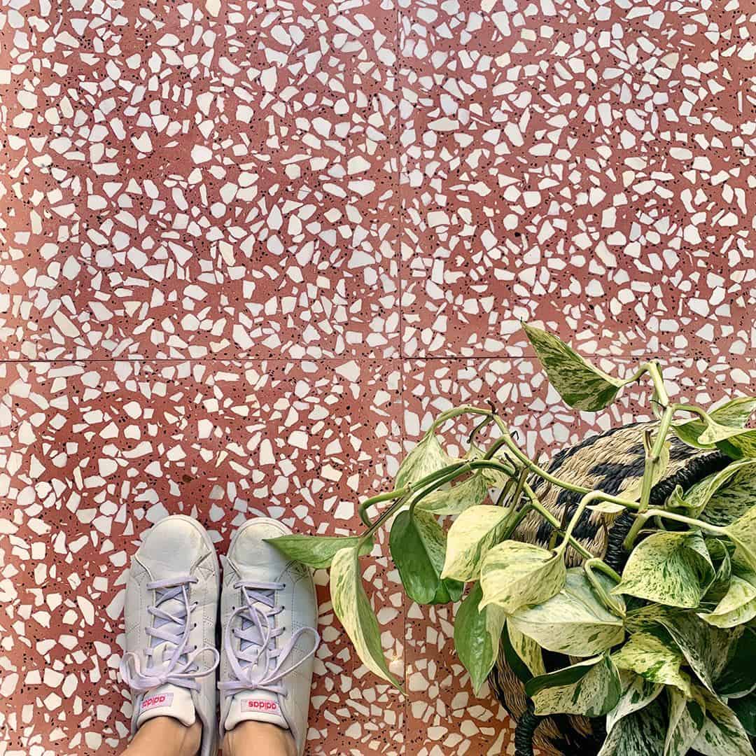 Person wearing white sneakers standing on a pink terrazzo tile floor beside a leafy potted plant.