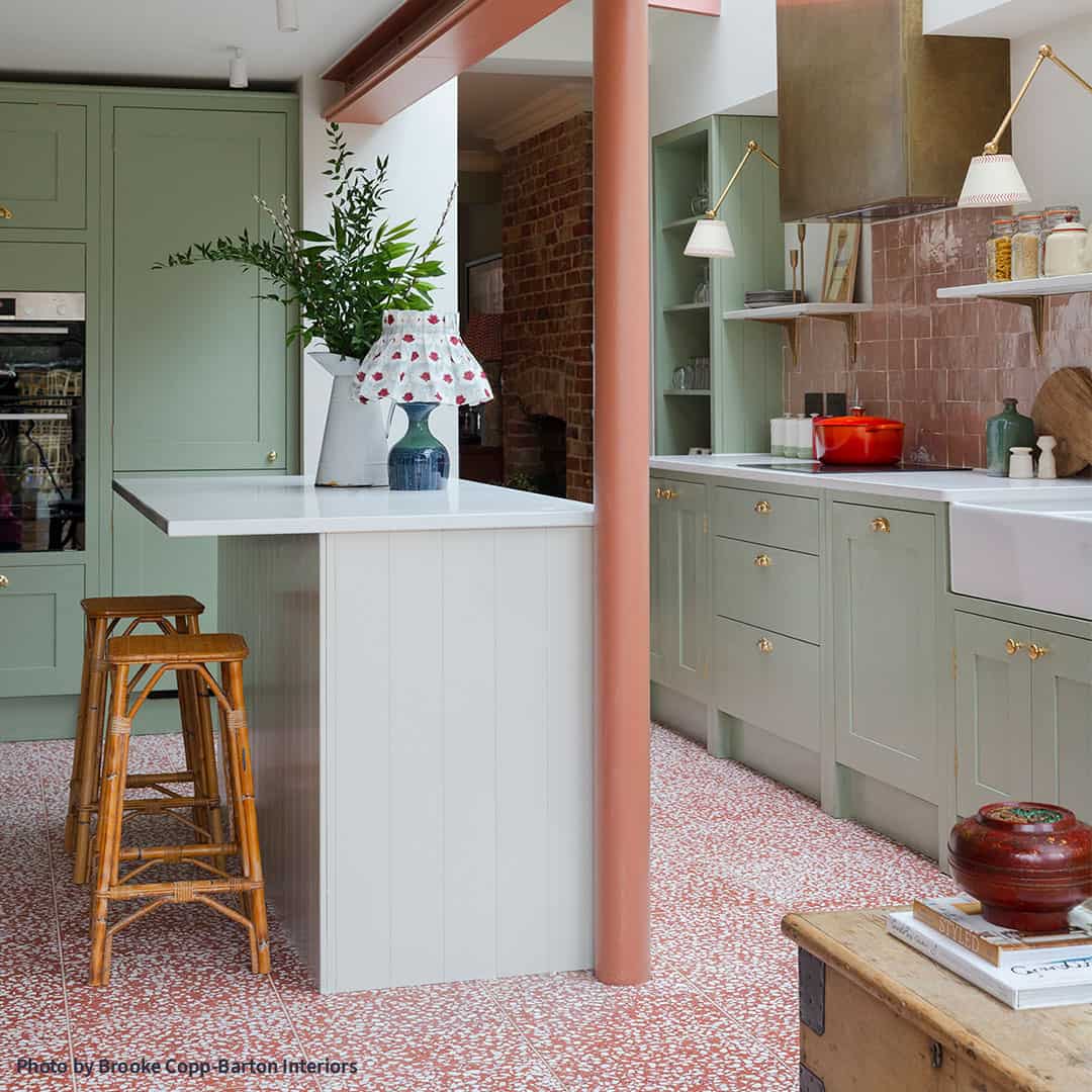 Modern kitchen with light green cabinets, white island with wooden stools, pink tile backsplash, brass fixtures, terrazzo floor, coral column, and exposed brick wall.
