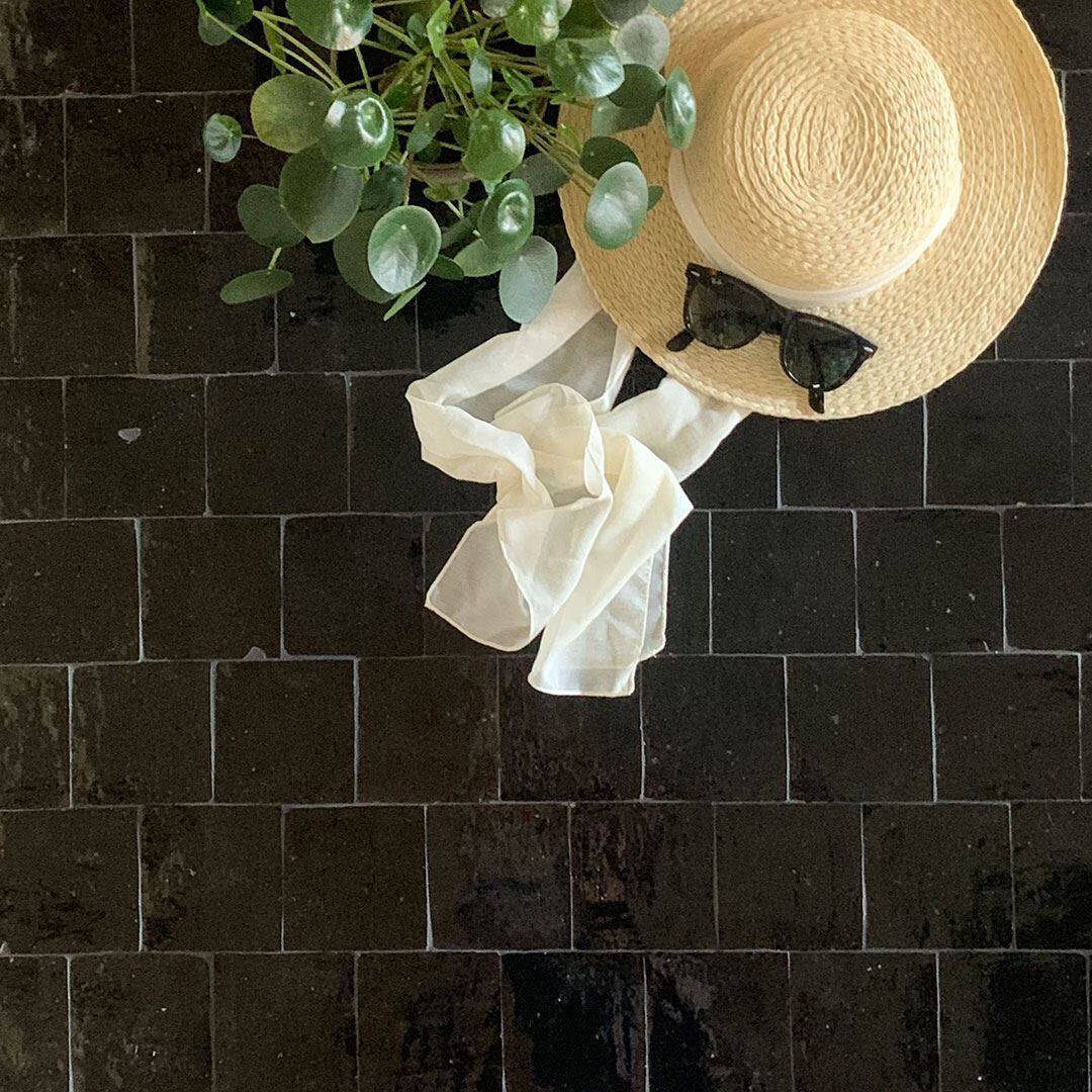 Flat lay with straw hat and white ribbon, black sunglasses resting on top, and potted plant with round leaves on glossy dark tiled surface, evoking summer leisure theme.