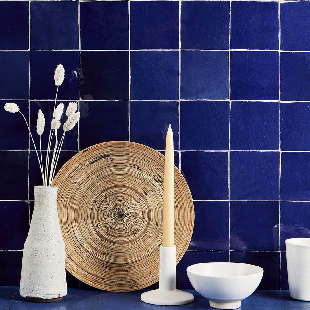 Minimalist decor with white vase, rattan plate, candle, and ceramics against glossy blue tiles.
