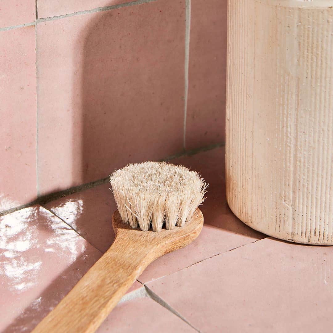 A close-up of a wooden bath brush with light bristles resting on glossy pink ceramic tiles.