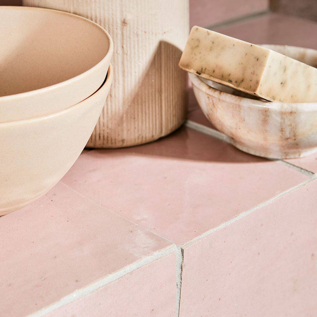 Stacked beige ceramic bowls, a ribbed cylindrical vase, and a small marble-like bowl holding a speckled soap bar, arranged on glossy pink tiles with visible grout lines.