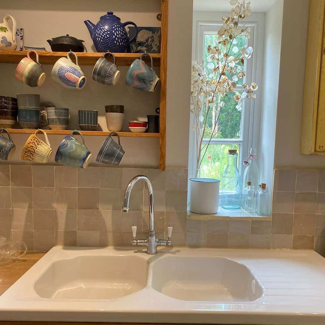 A cozy kitchen with a white double-basin sink beneath a small window. Glass bottles and a white pot with delicate dried stems sit on the windowsill. 