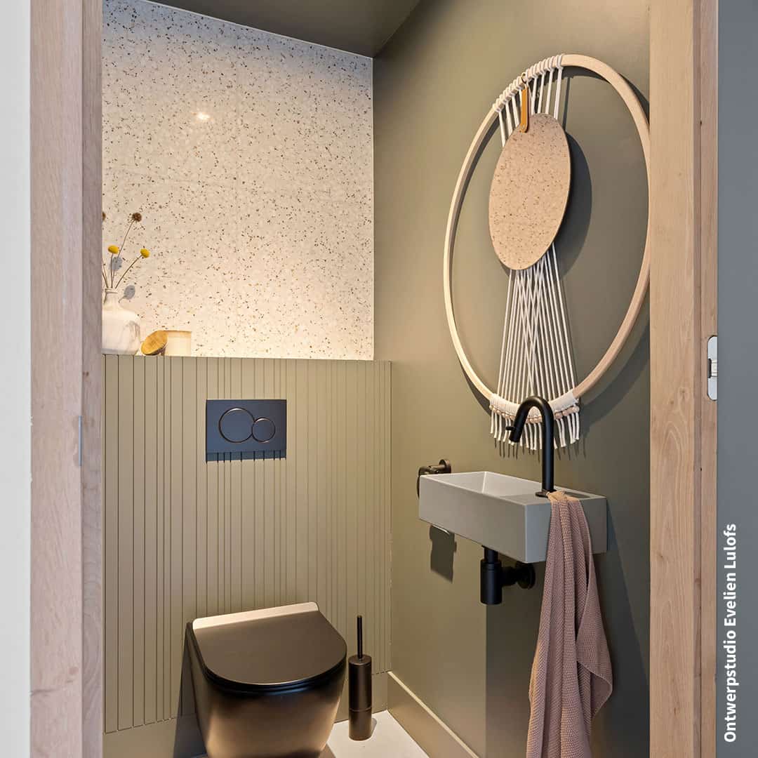 Small modern bathroom with matte black toilet, compact sink with black faucet, muted green paneled wall, terrazzo backsplash, and circular wall decoration above the sink.