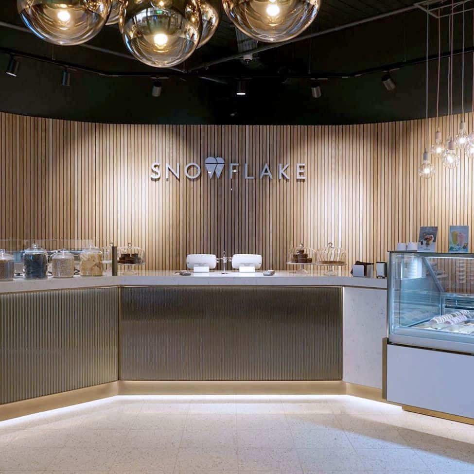 Modern dessert shop interior with “SNOWFLAKE” sign, ribbed metallic counter, glass jars of toppings, and gelato display case under warm lighting.