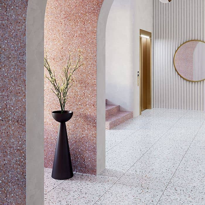 Modern interior with pink terrazzo archway, terrazzo staircase, white speckled floor, tall vase with branches, and gold-framed mirror beside a gold elevator door