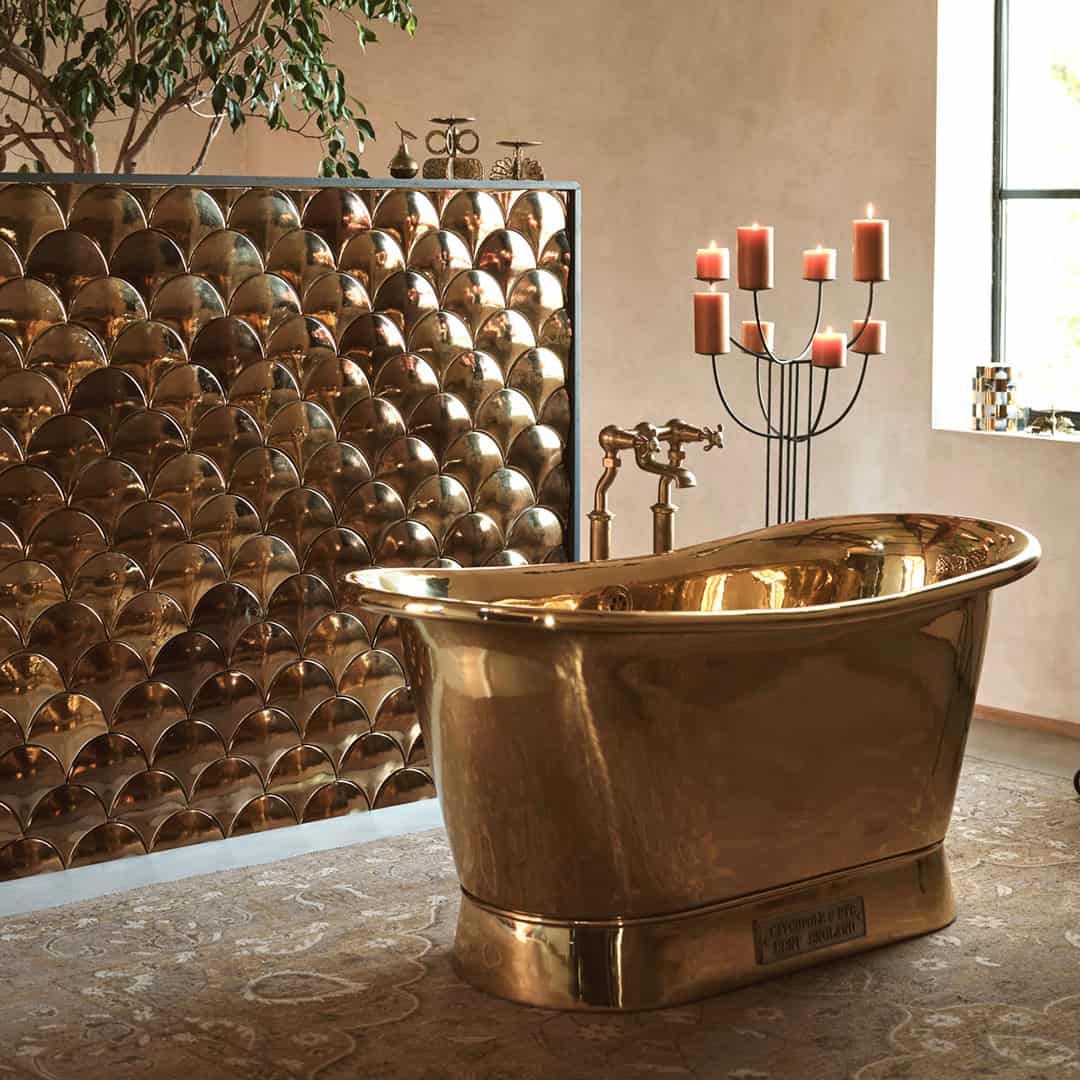 Luxurious bathroom with gold freestanding bathtub, metallic fish-scale tile wall, and candle stand beside window.