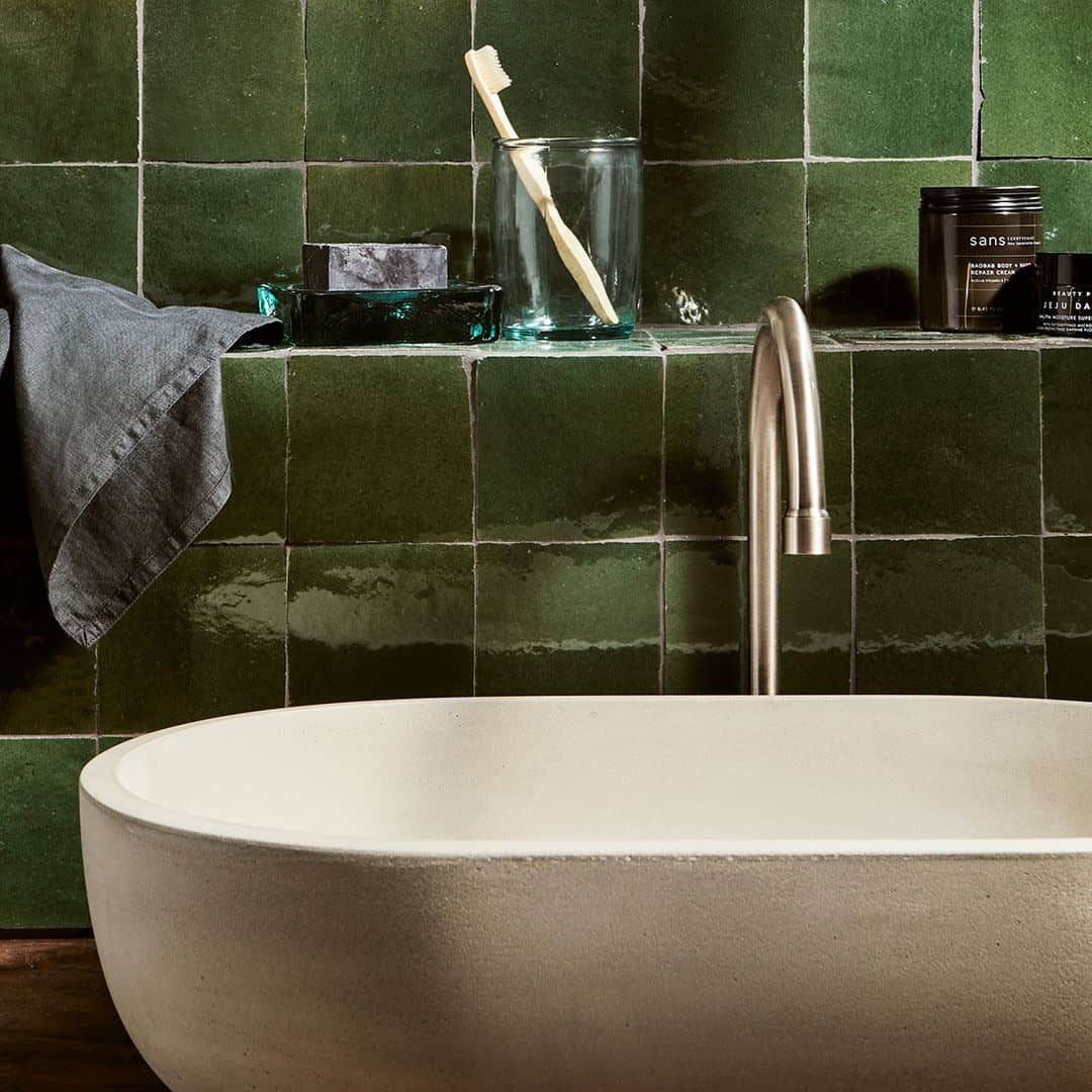 Close-up of a green tiled bathroom wall with a modern bathtub, shelf with glass jars, a toothbrush, and candles.