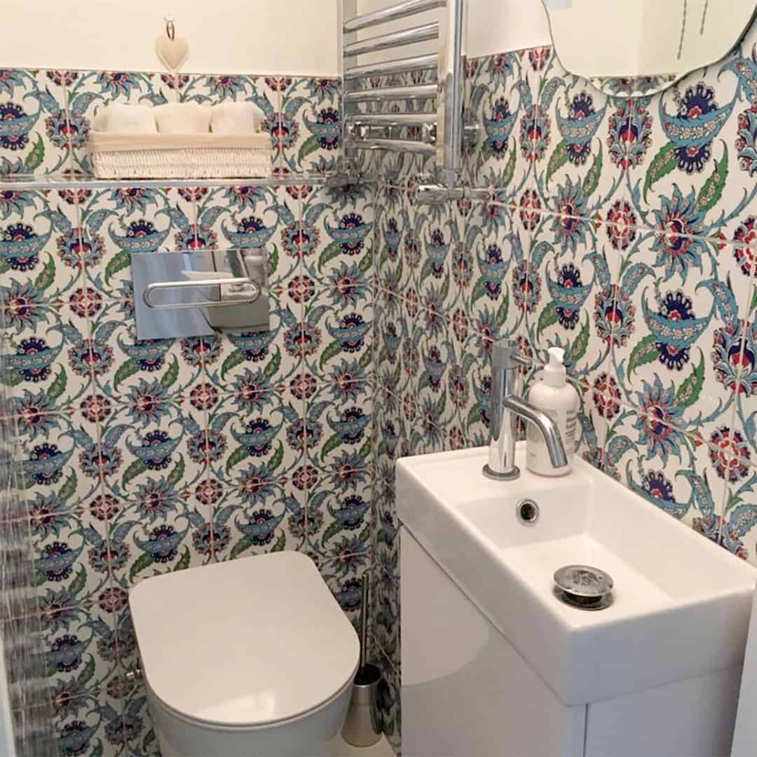 Compact bathroom with colorful patterned tile walls, wall-mounted toilet, small sink, chrome fixtures, and decorative shelf above.