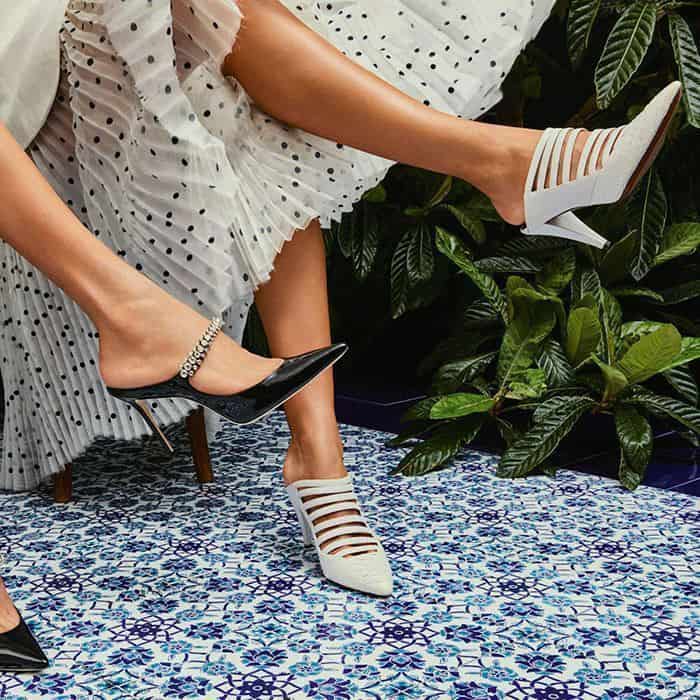 Two legs in polka-dot skirt wearing contrasting stiletto heels—one black glossy with embellished strap, the other white with cutout design—set against leafy plants and patterned blue-and-white tiled floor