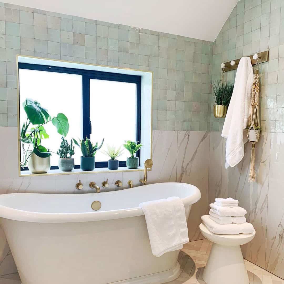 Elegant bathroom with a freestanding tub, gold fixtures, green tiled walls, and a window sill lined with potted plants.