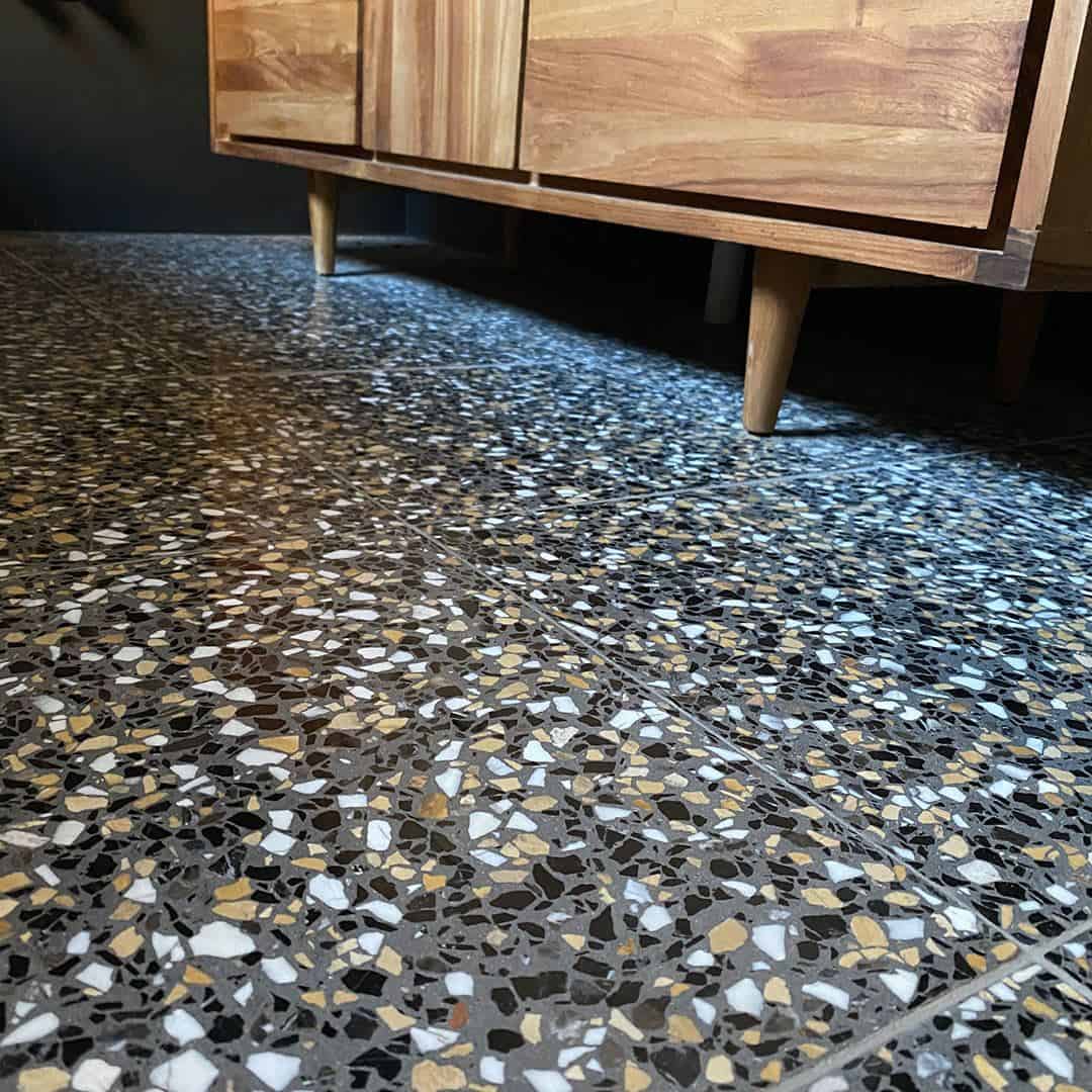 Dark terrazzo tile floor beneath wooden cabinet with tapered legs, showing multicolor stone chips.