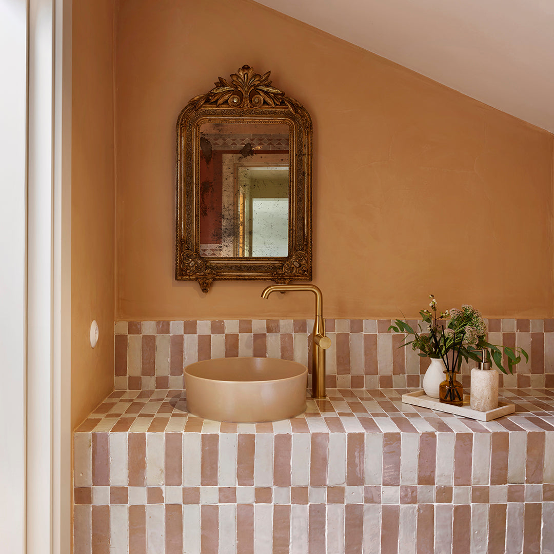 Elegant bathroom with a beige sink, gold faucet, and a vintage mirror, set against a wall of light pink and white striped tiles. There are decorative vases with flowers and candles on the countertop.