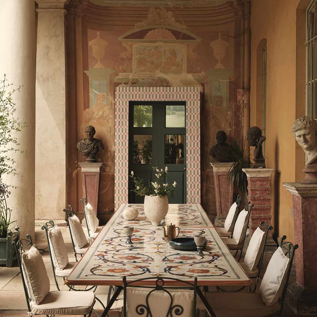 Elegant outdoor dining area with a large mosaic table, surrounded by chairs, classical busts, and a decorative green door framed with pink tiles. The background features frescoed walls.