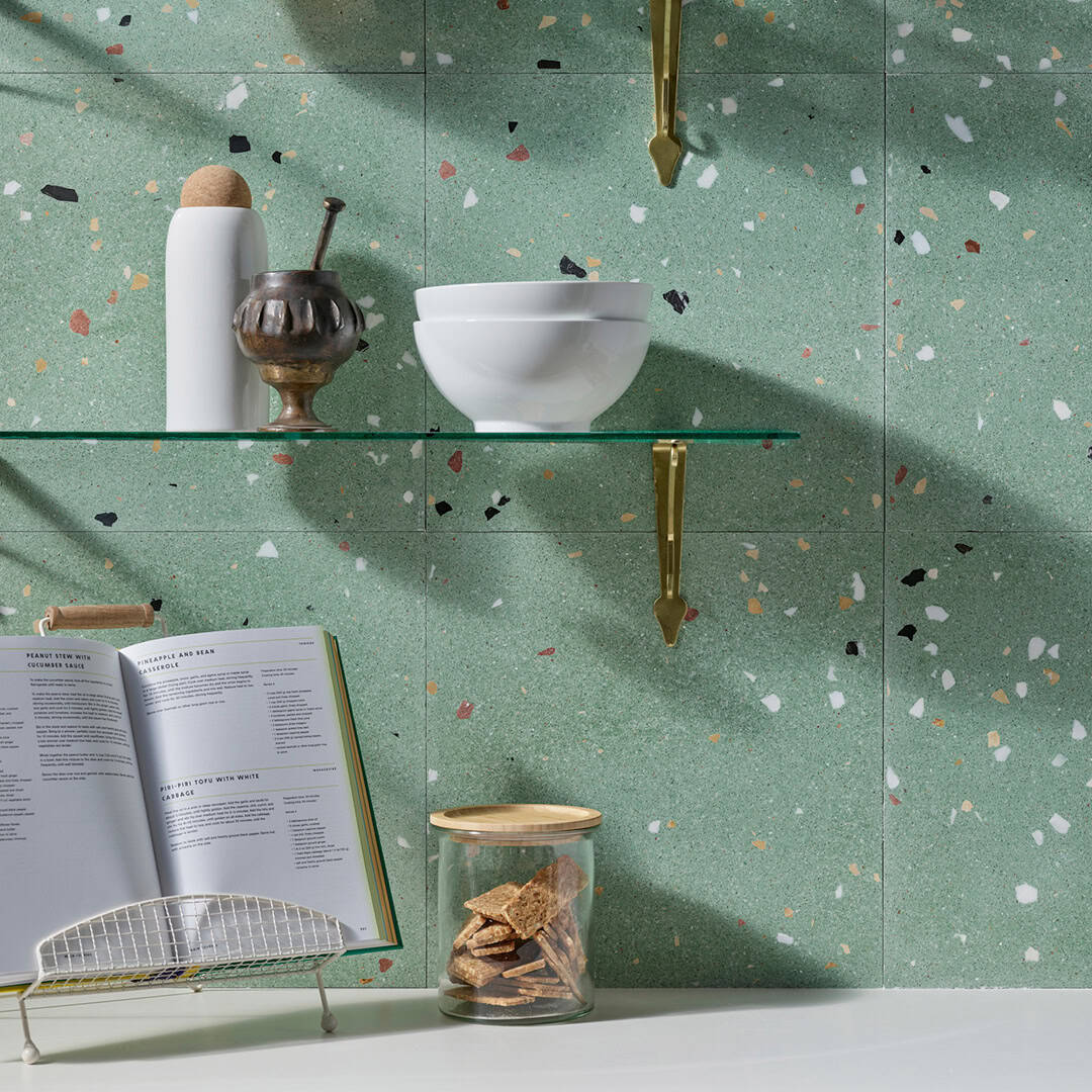 Kitchen wall with green terrazzo surface, brass shelf holding bottle, cup, and bowl, open cookbook on stand, and glass jar of crackers.