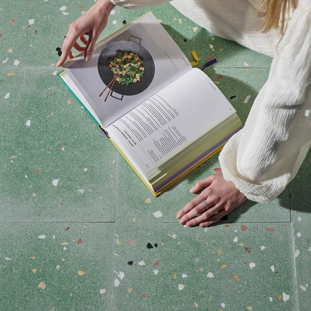 Person in white textured garment sitting on green terrazzo floor, reading an open cookbook with photo of stir-fry in black wok and recipe text on facing page.