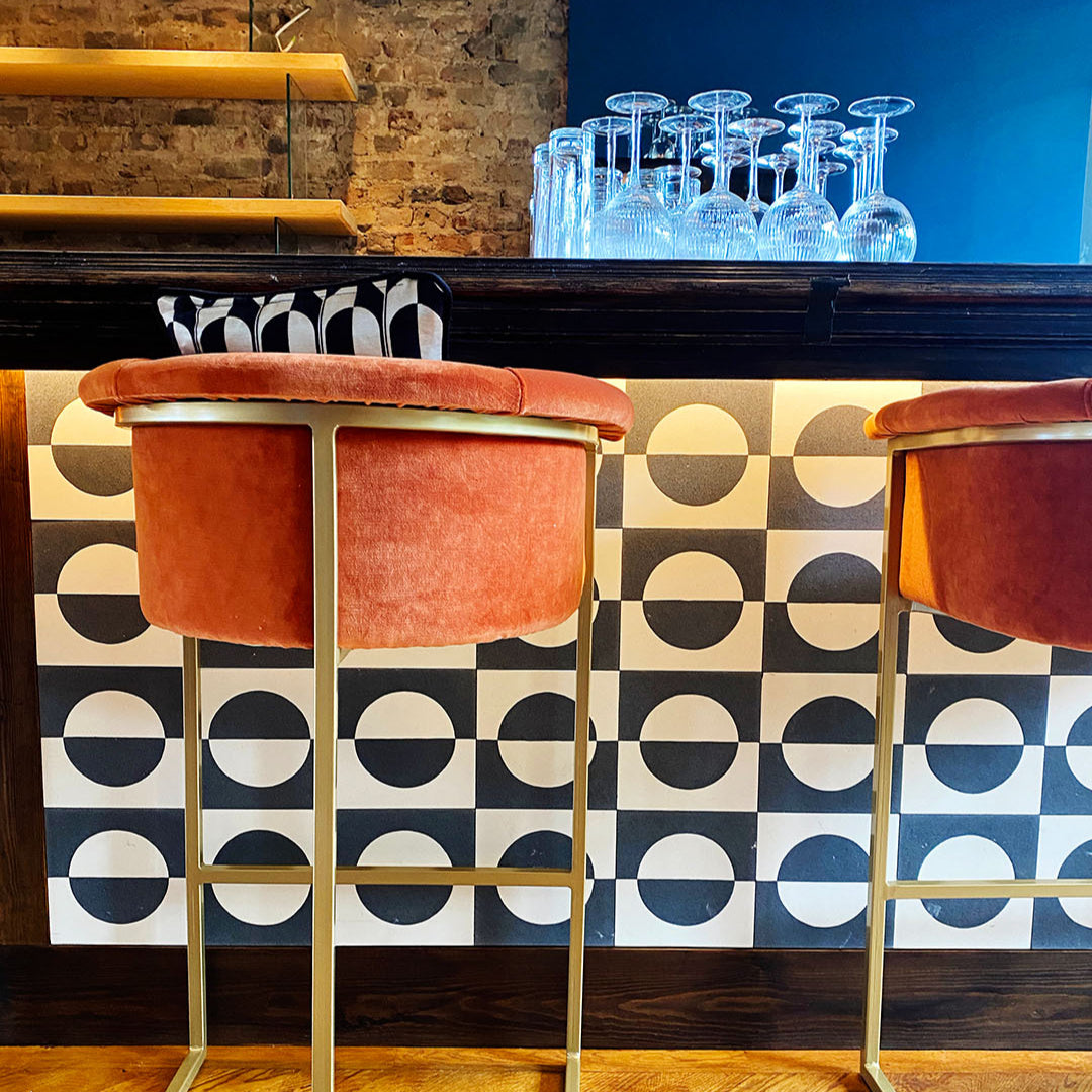 Bar stools with orange seats in front of a bar counter with glasses, against a geometric patterned wall.