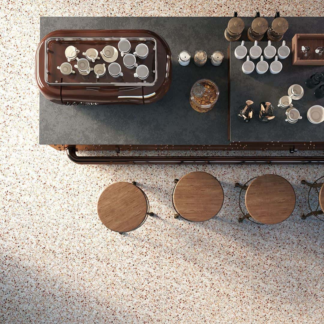 Overhead view of café counter with espresso machine, cups, wooden stools, and terrazzo flooring in warm tones.