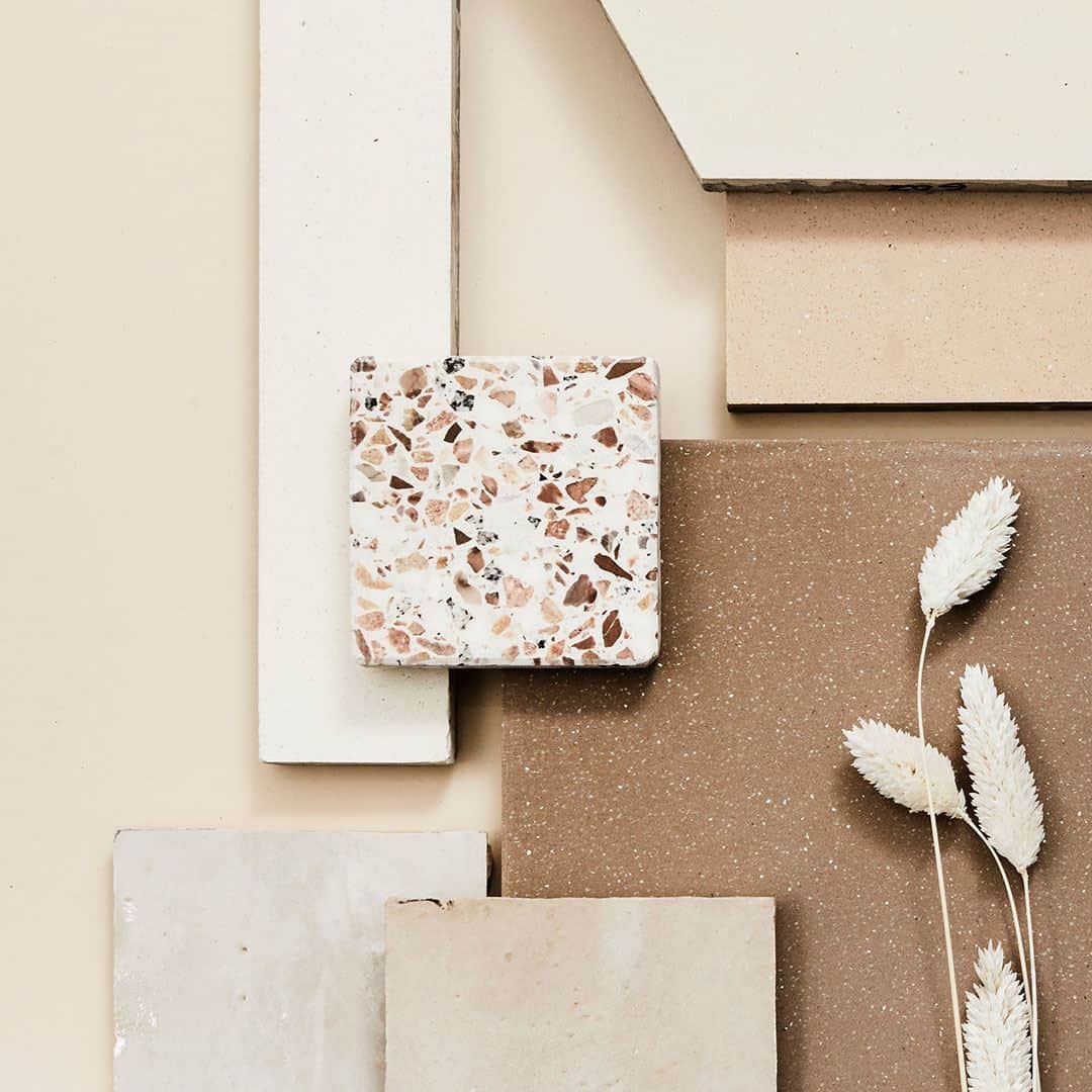 Layered neutral tile samples with terrazzo square and beige stone slabs, styled with dried grass accents.
