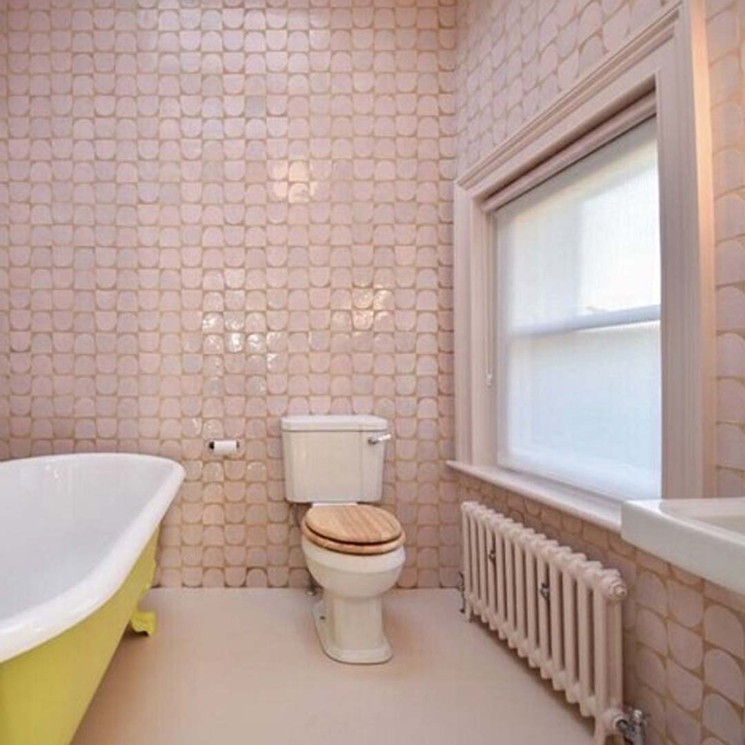 A bathroom interior with a white toilet featuring a wooden seat, a yellow clawfoot bathtub partially visible to the left, and a white radiator beneath a frosted window. 
