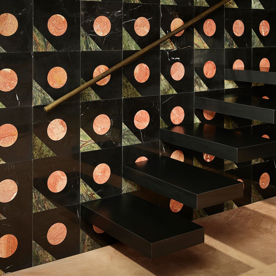 Modern staircase with floating black steps against tiled wall featuring large pink-orange circles on black background with green marble accents, and sleek metallic handrail.