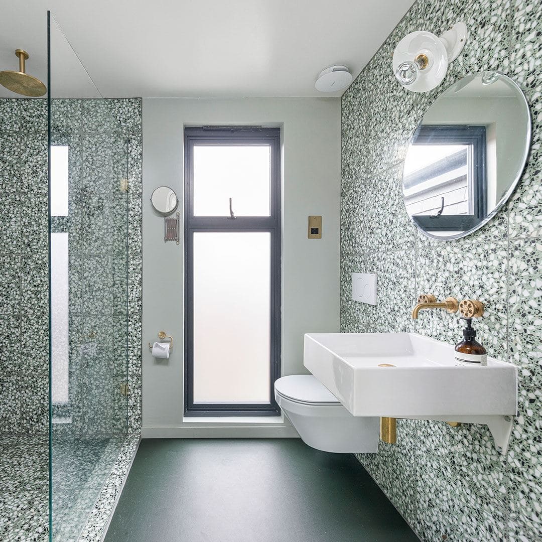 Bright bathroom with green terrazzo walls, glass shower, wall-mounted sink, brass fixtures, and frosted window.