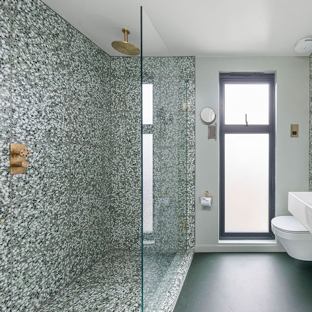 Bright bathroom with green terrazzo walls, glass shower, wall-mounted sink, brass fixtures, and frosted window.