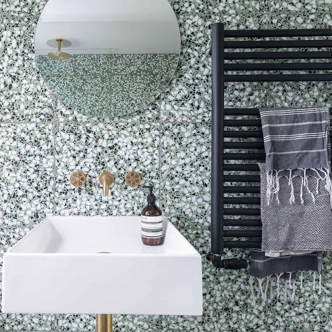 Bathroom with green terrazzo wall tiles, white sink, round mirror, brass fixtures, and black towel radiator.