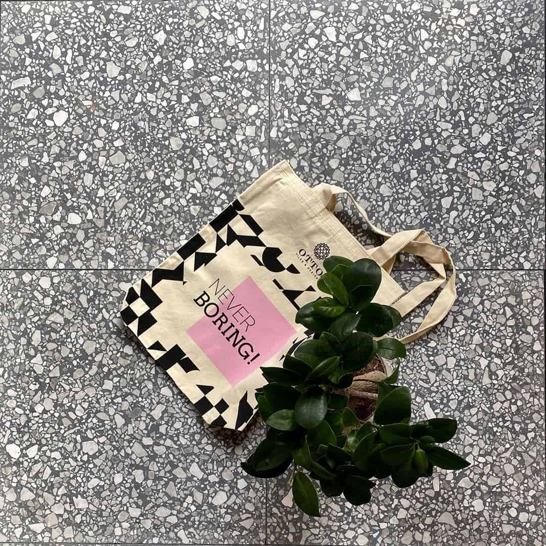 Canvas tote bag with plant on gray terrazzo floor, featuring graphic text and geometric black pattern design.
