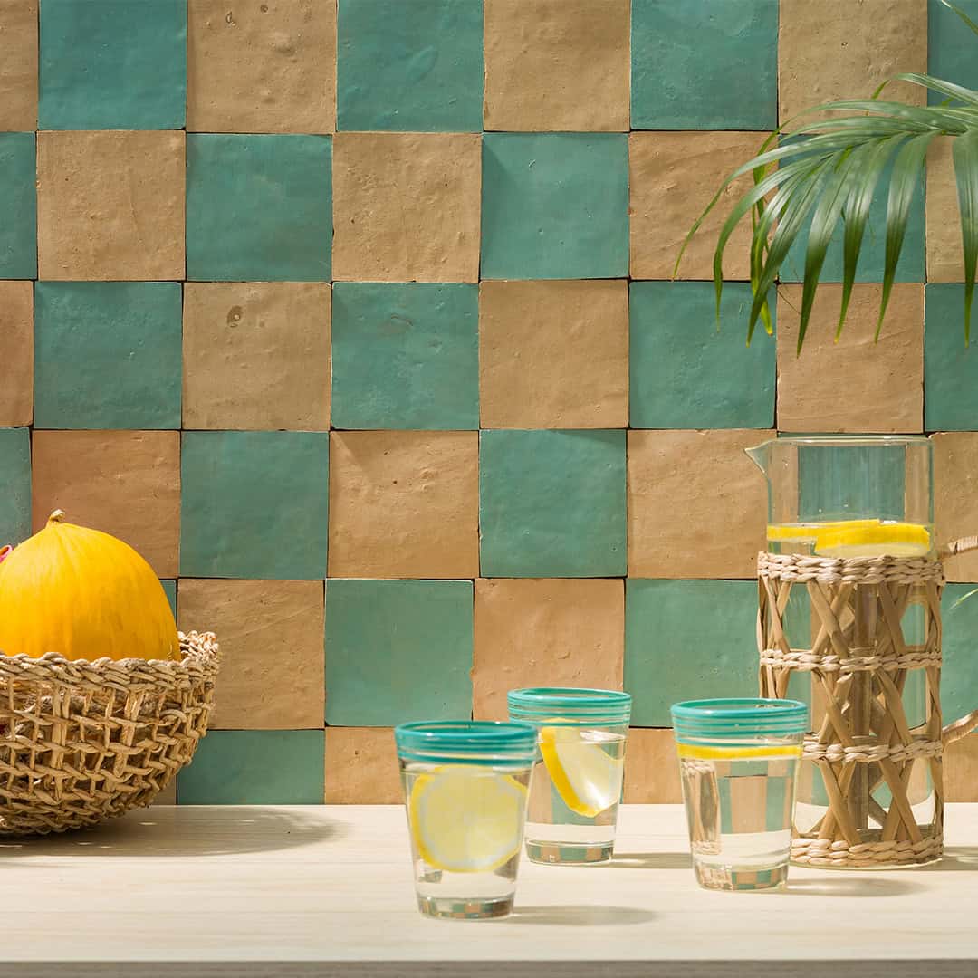 Tabletop scene with turquoise and beige checkered tiles, woven basket holding yellow melon, three glasses of lemon water, decorative pitcher, and palm leaf creating a refreshing summer atmosphere.