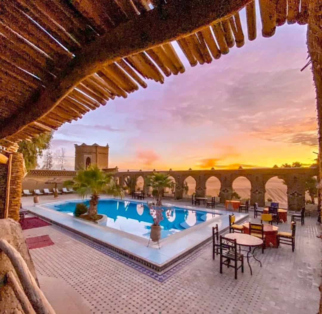 The pool is framed by vibrant palm trees and lounge chairs, with an arched structure in the distance adding an architectural touch. The floor is covered with intricate tiled patterns, adding a traditional, colorful flair to the scene.