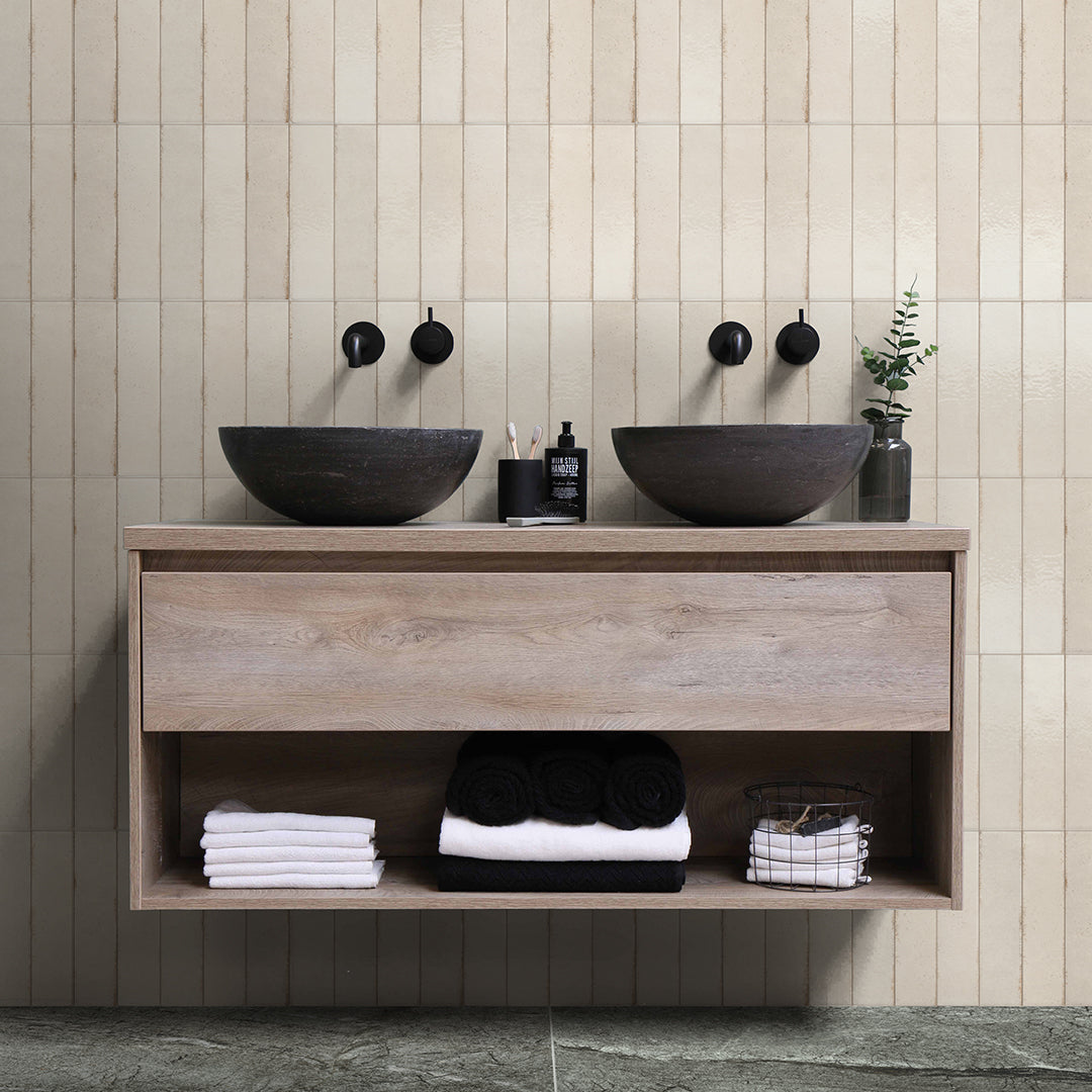 Bathroom vanity with two dark stone sinks, wooden cabinet, and folded towels on a tiled wall background.