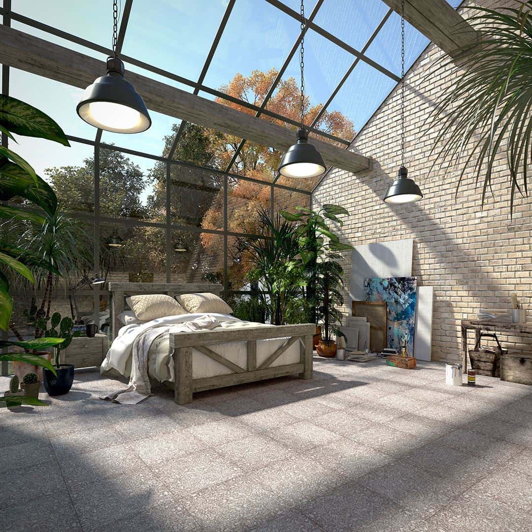Sunlit bedroom with glass ceiling, exposed brick walls, indoor plants, and neutral terrazzo tile flooring.