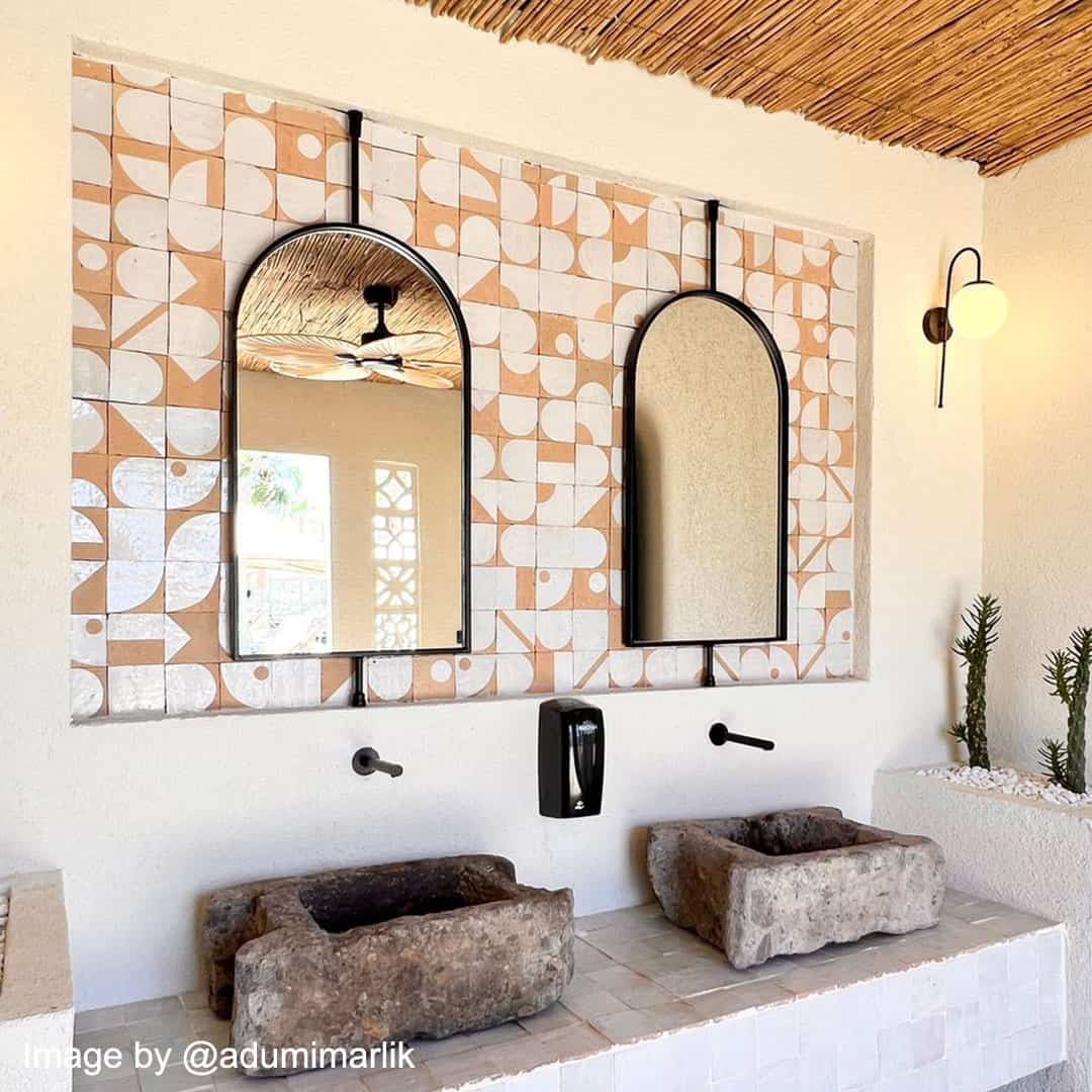 Chic bathroom with a geometric tile wall in terracotta and white, featuring two arched mirrors, stone sinks, and minimalist faucets.