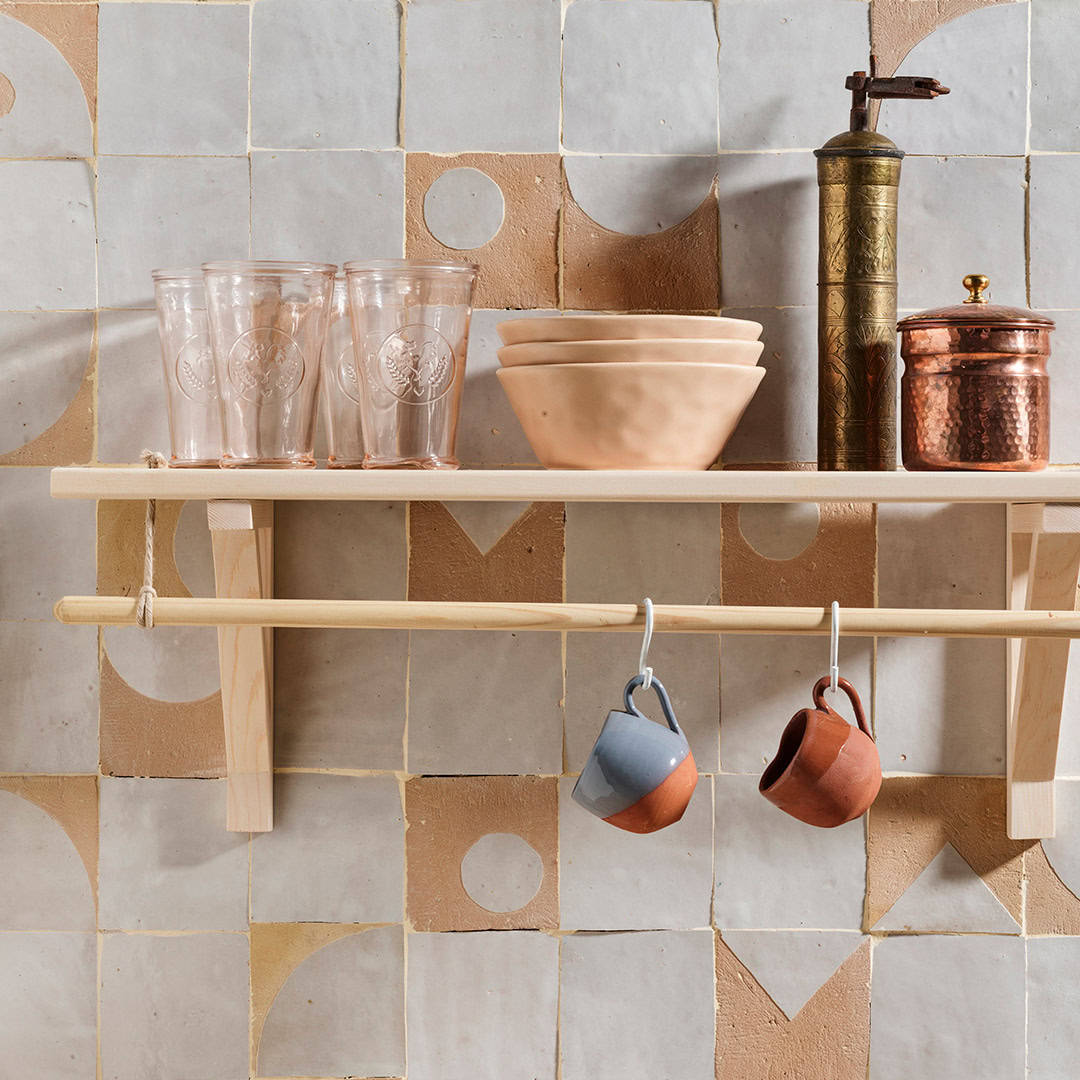 Wooden shelf with glass cups, terracotta bowls, a vintage coffee grinder, and hanging mugs against a geometric tile wall in earthy tones.