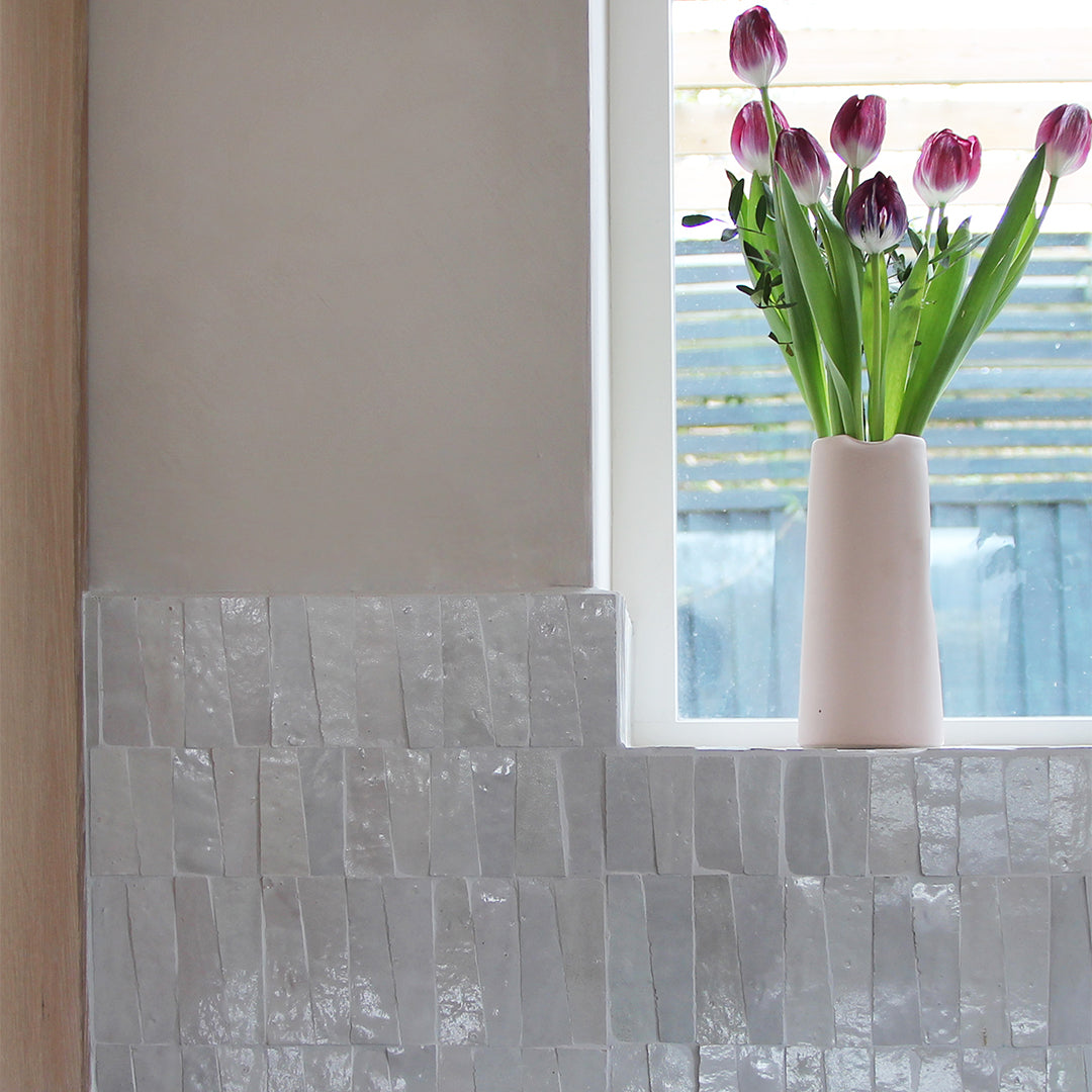 Vase with purple tulips on a tiled wall next to a window