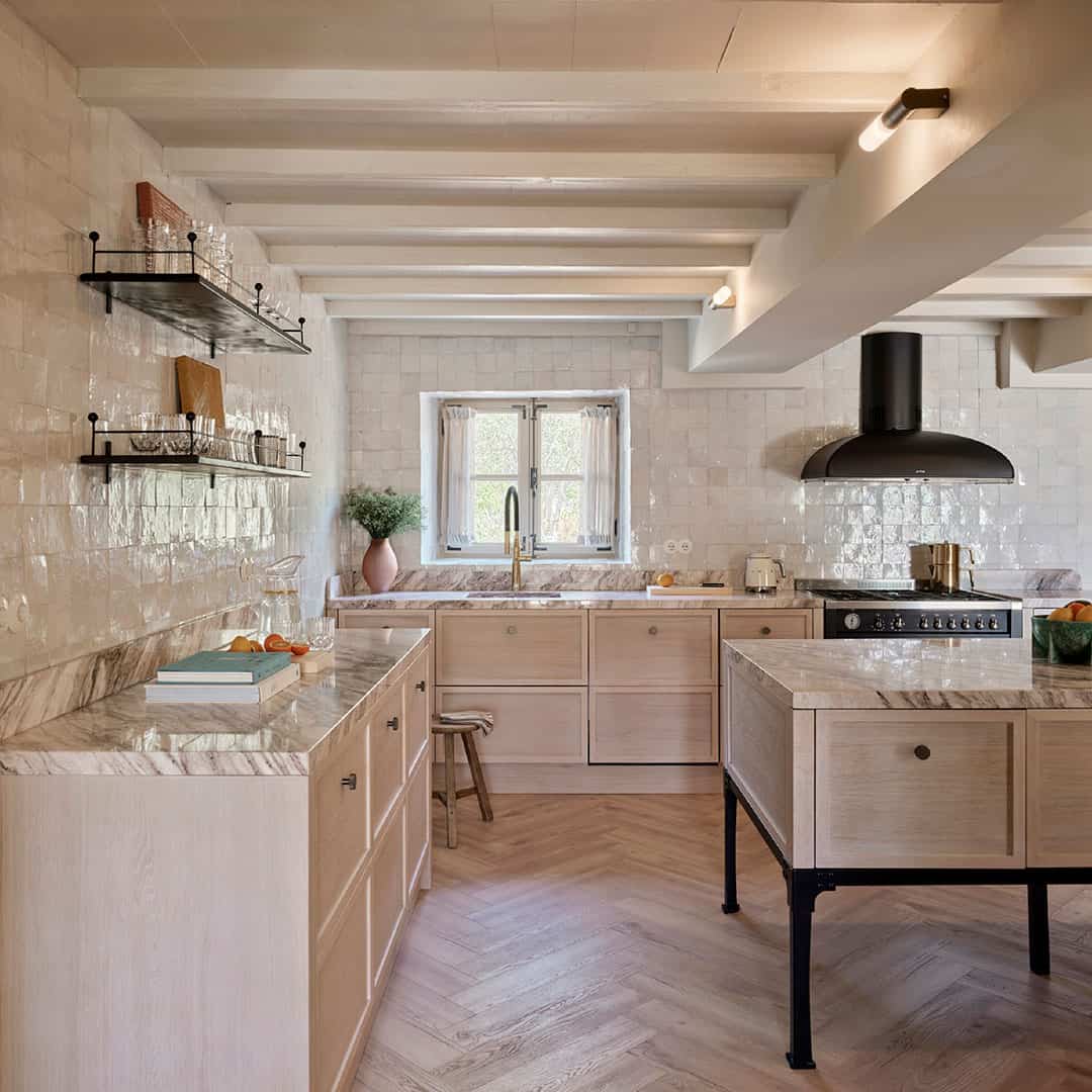 A modern rustic kitchen with light wood cabinets, marble countertops, and a herringbone wooden floor. 