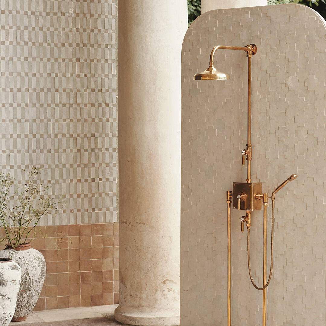 Elegant copper shower fixture with rainfall head and handheld sprayer against textured neutral tile wall.